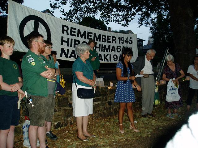 Banner: Remember 1945. Lady in blue dress with long hair speaks on the river bank