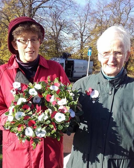 Two ladies holding a wreath of white poppies against a background of bare trees in the sun