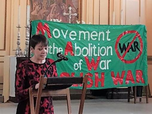 Lady with short hair and a red and black dress reading a lecture, in front of an altar with silver candlesticks