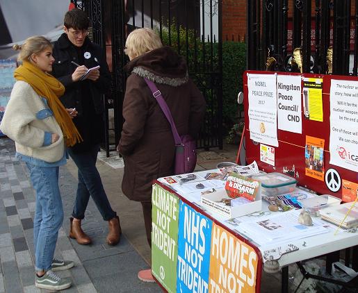 three people standing out of doors at a table covered in leaflets, with a white cardboard tray of white poppies, saying 'Remember and Disarm' and 'wear a white poppy for peace'