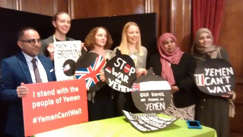 six people standing in a panelled room holding placards reading 'Our bombs are falling on Yemen', 'I stand with the people of Yemen', 'Yemen can't wait' etc
