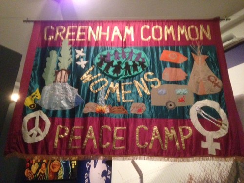 banner with outlines of women, a carvan, wildlife etc and the words 'Greenham Common Womens Peace Camp'