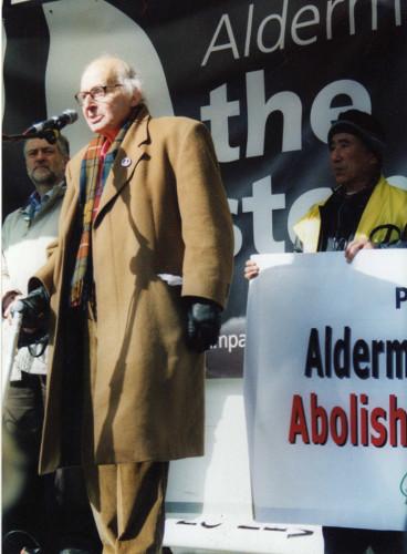 man in a brown overcoat standing and speaking at a microphone
