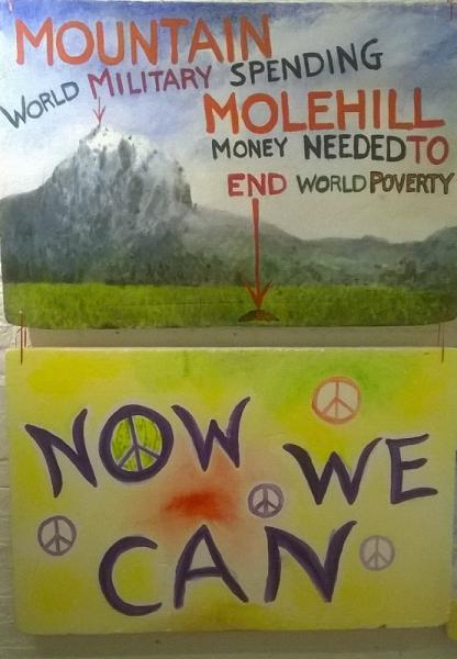 more hand-painted placards: 'drawings of a mountain (world military spending) and a molehill (money needed to end world poverty)' and a slogan 'Now we can' with CND logos