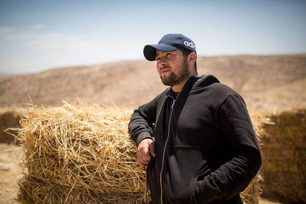 a bearded man leans on a bale of straw or hay in the sunshine