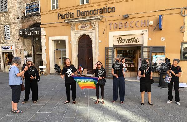 A row of six women in black with placards and a rainbow PACE (peace) flag